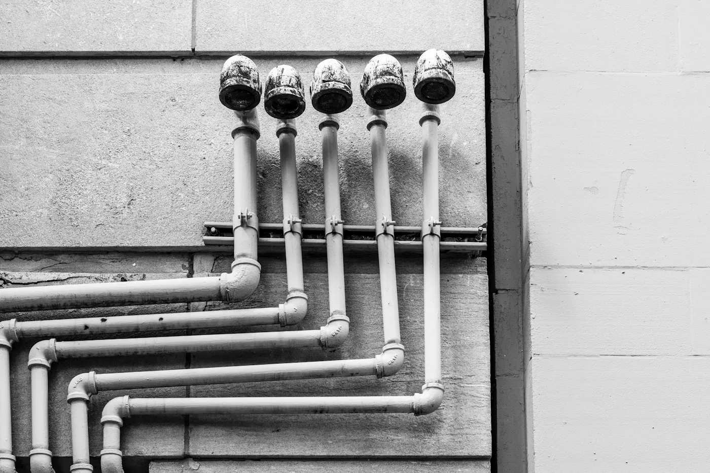 Cinq colonnes de tube acier peint montant le long d’un mur de parpaing, coudées à leur base et coiffées d’un chapeau grillagé