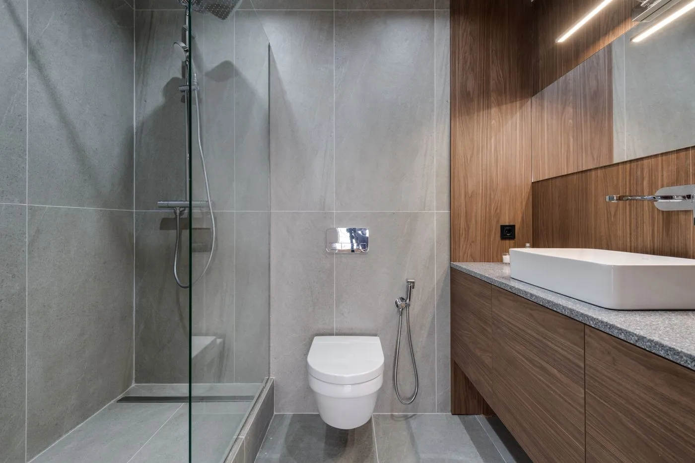 Salle de bains carrelée en grès gris, douche de plain-pied à paroi vitrée et meuble en noyer