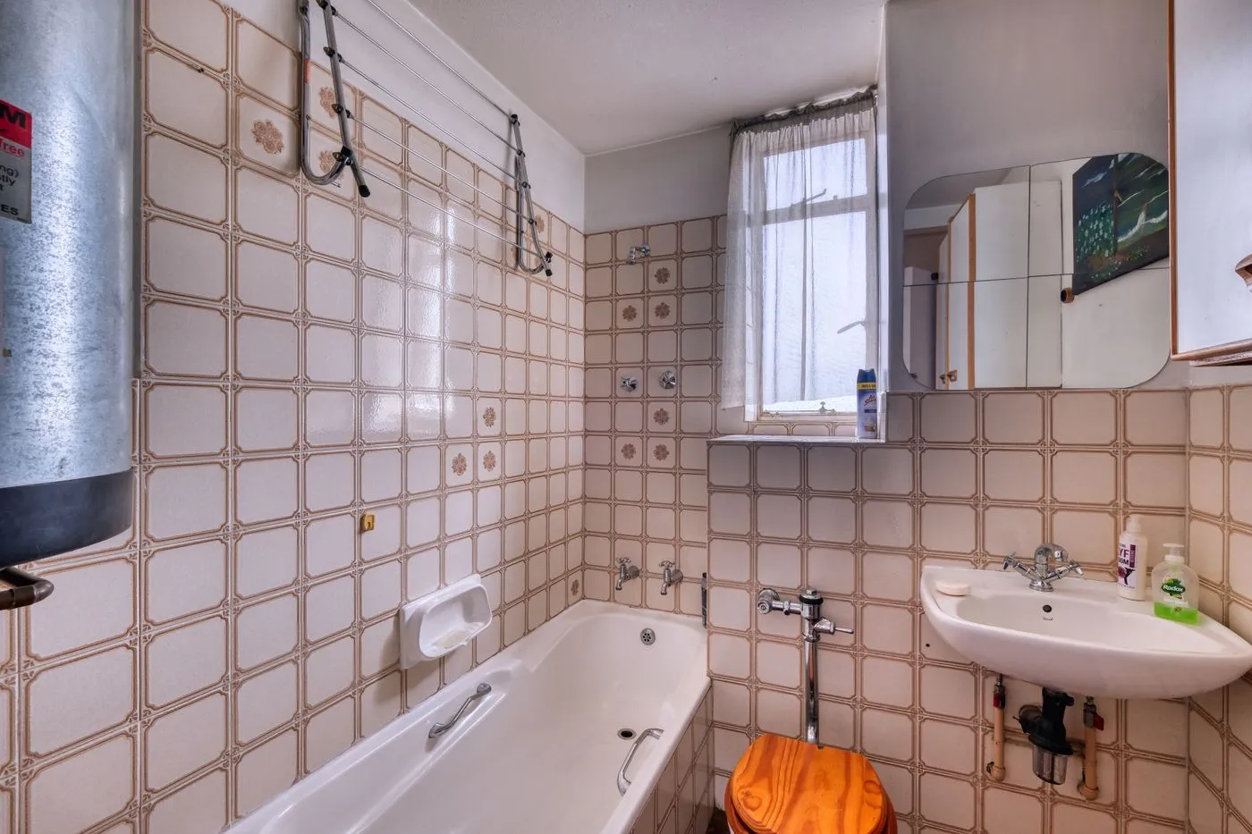 Salle de bains d’origine : faïence beige à motifs, baignoire encastrée, chauffe-eau électrique accroché au-dessus